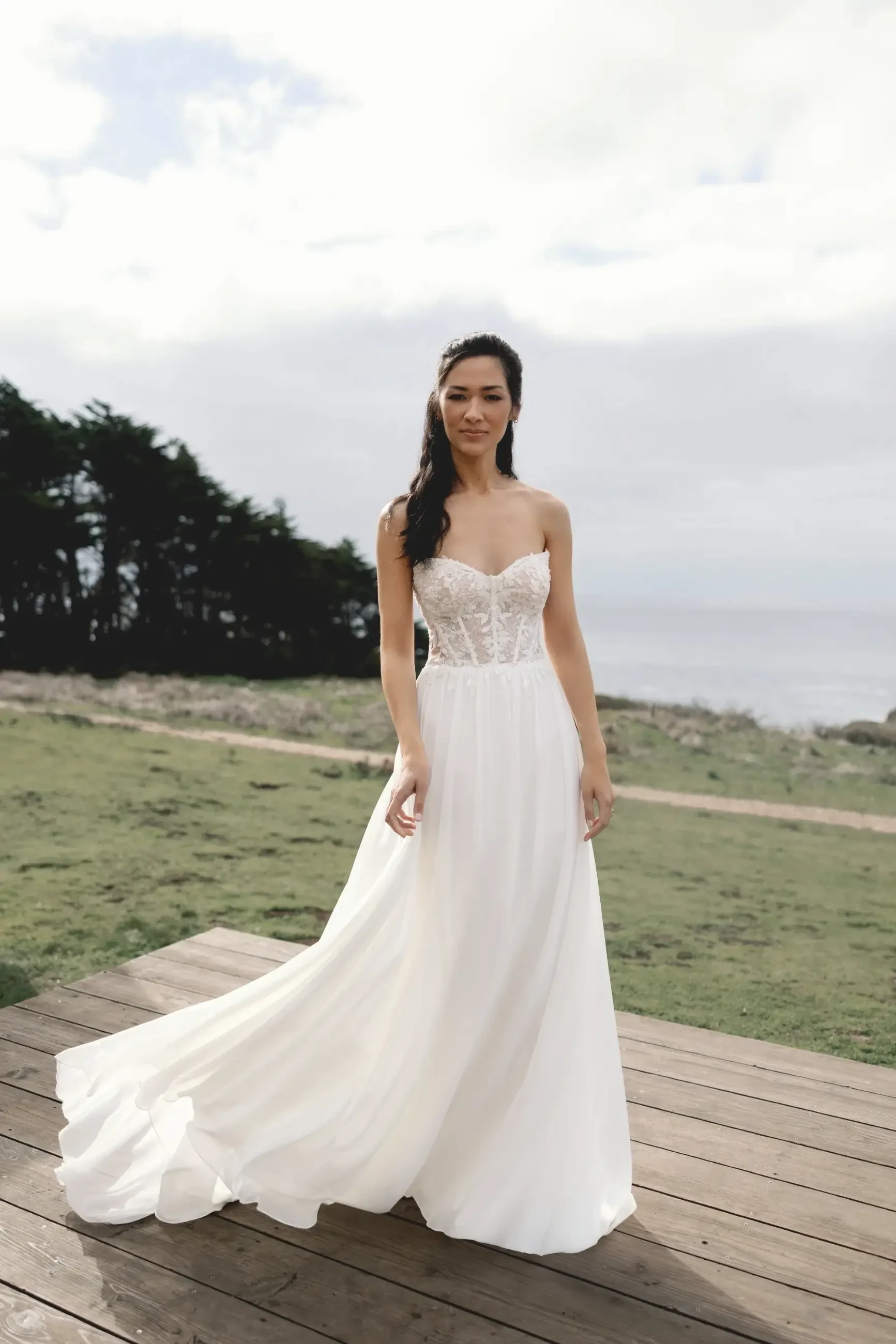 A woman in a strapless white wedding gown stands on a wooden deck with green grass and trees in the background, under a cloudy sky.