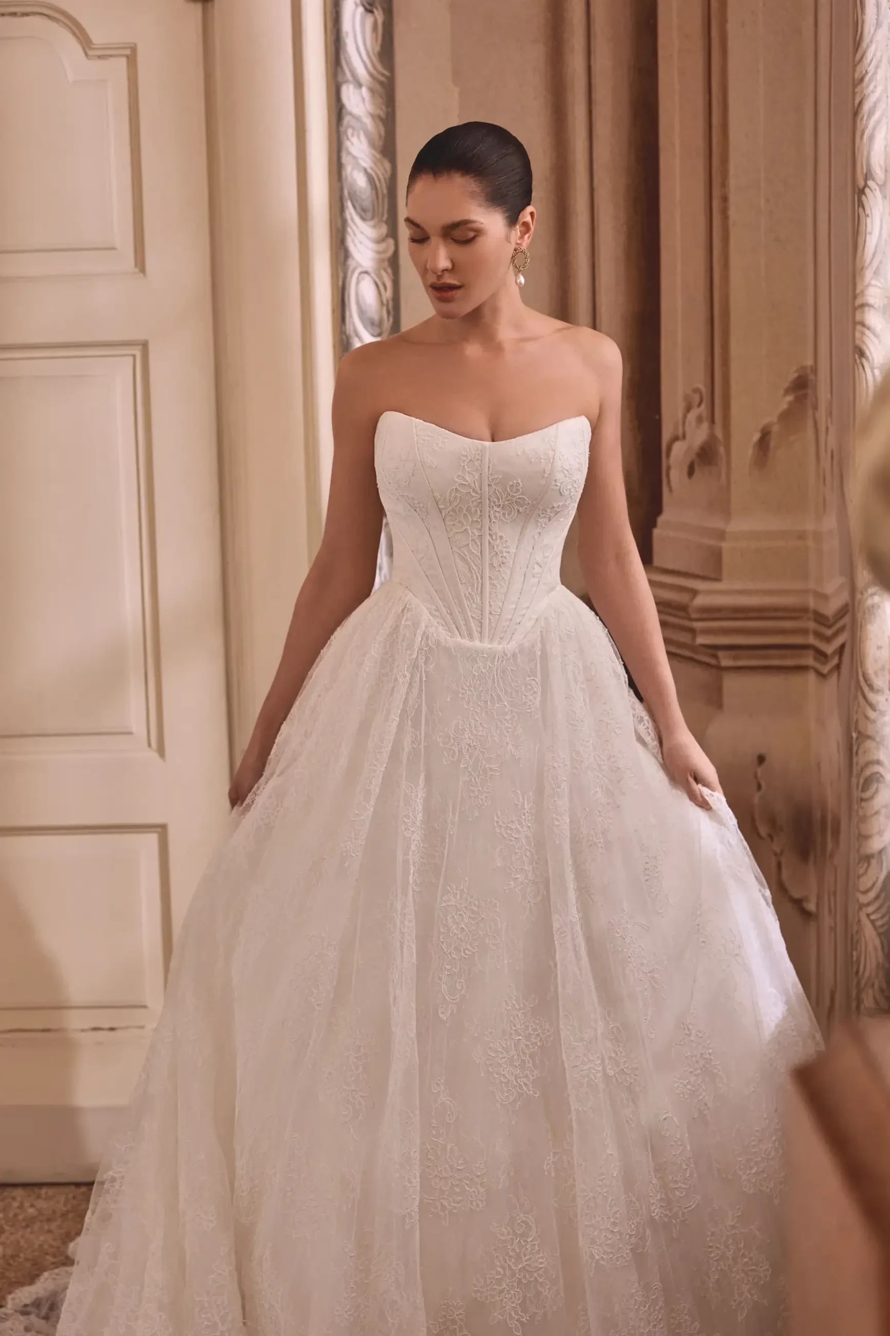 A woman wearing a strapless white wedding dress with a full skirt, standing in an elegant interior space.