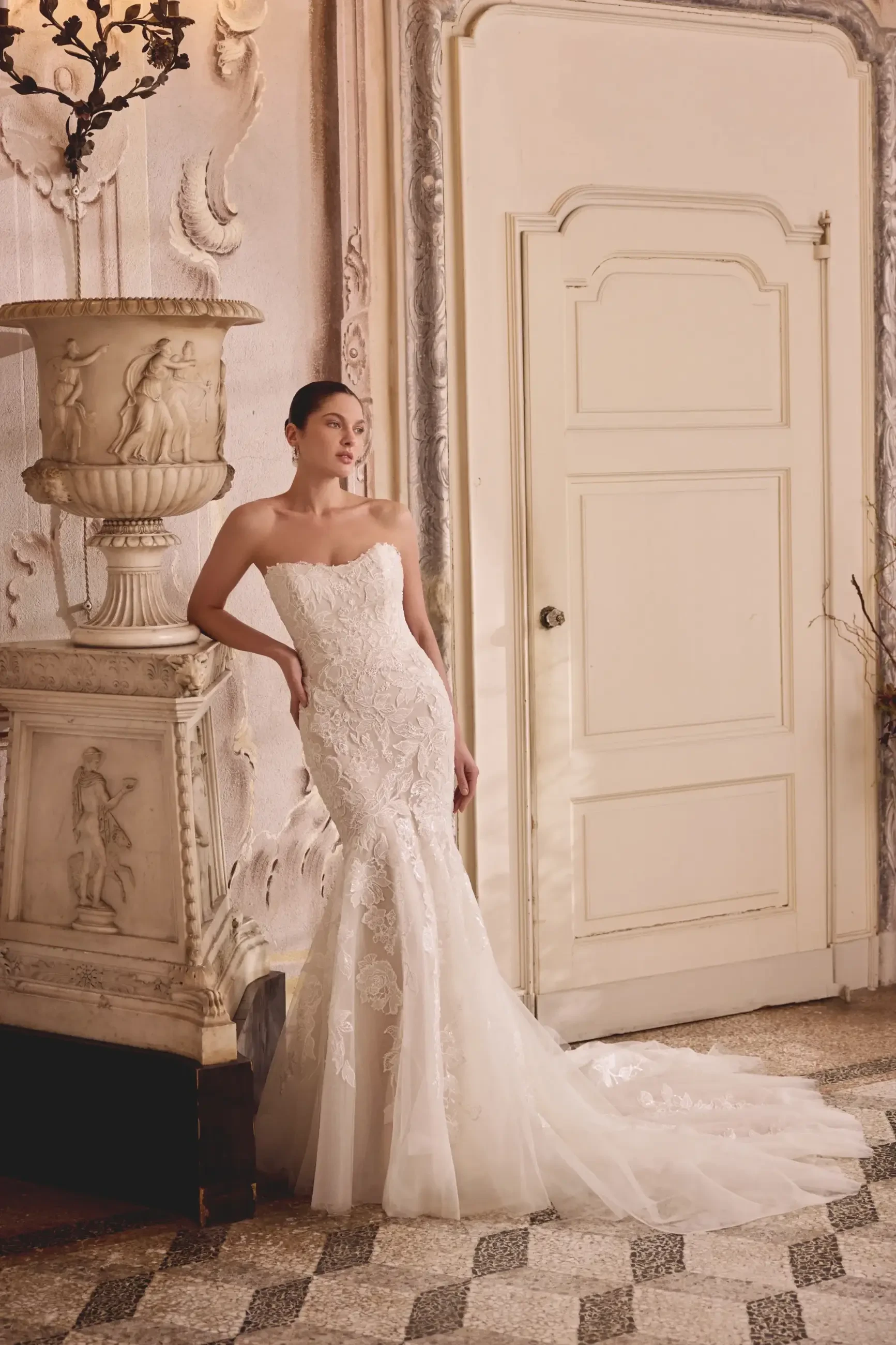 A woman in a strapless wedding dress with intricate lace detailing poses next to an ornate vase and door in a stylish interior setting.